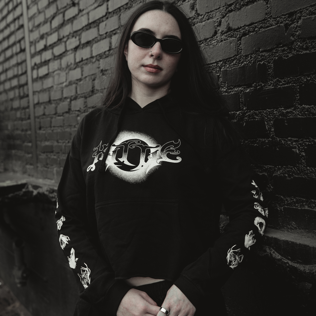 A person with long dark hair and black sunglasses wears a Kittie women's cropped hoodie made from ringspun cotton, featuring white graphics, while standing against a brick wall and looking at the camera with a neutral expression.
