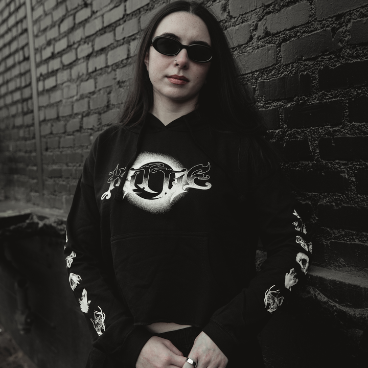 A person with long dark hair and black sunglasses wears a Kittie women's cropped hoodie made from ringspun cotton, featuring white graphics, while standing against a brick wall and looking at the camera with a neutral expression.