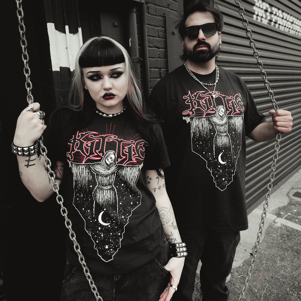 Two people with dark makeup and gothic style stand outside in Kittie garment-dyed Comfort Colors T-Shirts featuring a red and white occult design. The woman holds a chain, while the man stands confidently behind her in bold ring-spun cotton shirts.