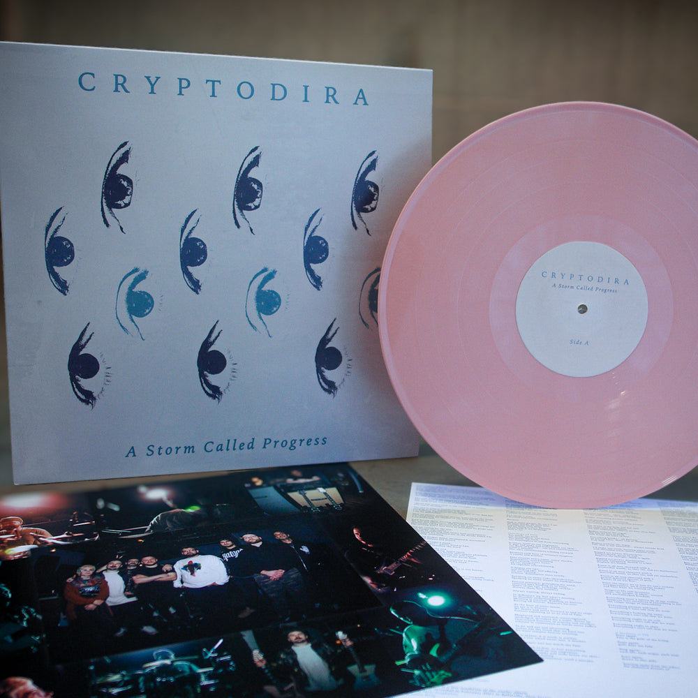 A limited edition pale pink Cryptodira vinyl sits beside its album cover—featuring stylized blue eyes and "Cryptodira: A Storm Called Progress"—with an insert of band photos and a lyric sheet placed below.