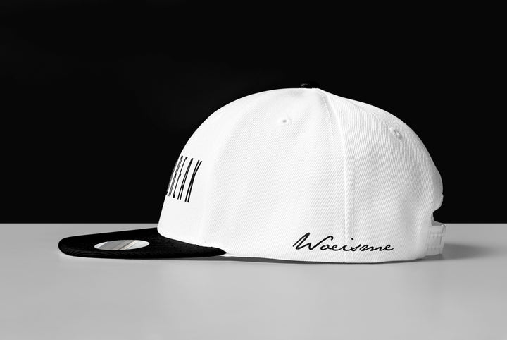 A white and black Woe Is Me baseball cap rests on a white surface against a black background. Perfect for Daybreak album fans, it features a black brim and the word Wolverine in script on the side.