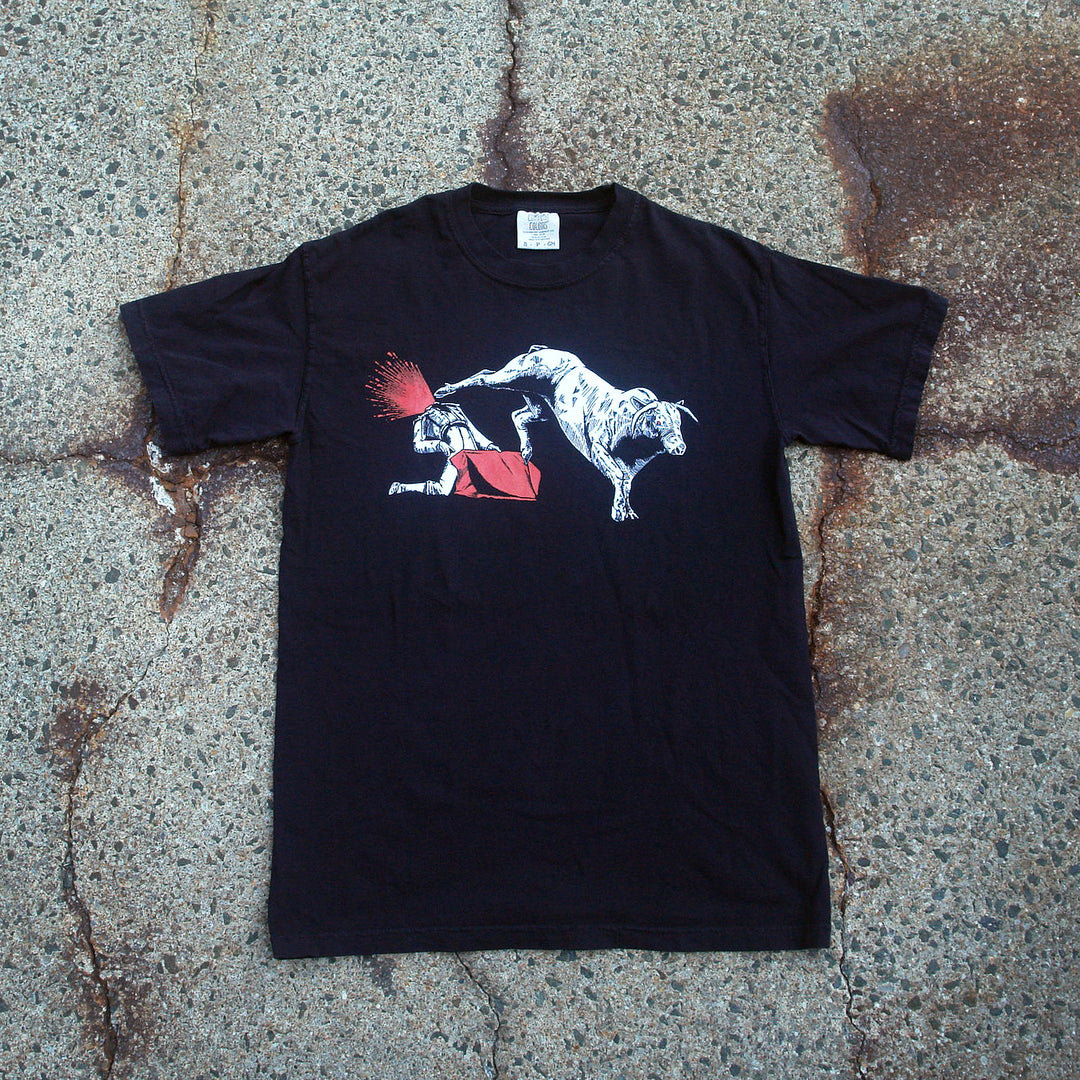 MerchNow Black Comfort Colors T-Shirt, made of soft garment-dyed ring-spun cotton, shown on rough concrete. Features a bold white dinosaur kicking a person’s head, with red highlighting an explosive impact.