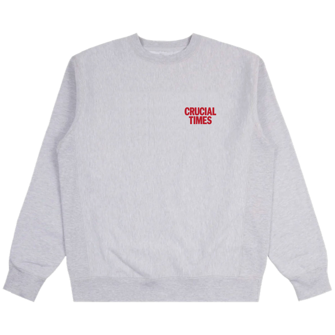 A light gray crewneck sweatshirt from Crucial Times with bold red CRUCIAL TIMES print on the upper left chest. This classic piece features ribbed cuffs and hem. Shipping starts 2/20/26.