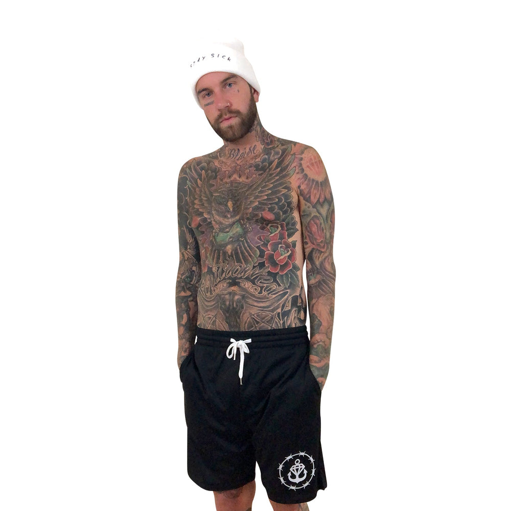 A bearded, heavily tattooed man poses shirtless in Stay Sick Clothing’s Black Court Shorts with hands in pockets, wearing a white beanie. He stands against a plain white background, showcasing the bold style of the Barbed Logo collection.
