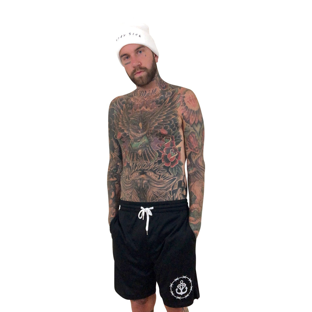 A bearded, heavily tattooed man poses shirtless in Stay Sick Clothing’s Black Court Shorts with hands in pockets, wearing a white beanie. He stands against a plain white background, showcasing the bold style of the Barbed Logo collection.