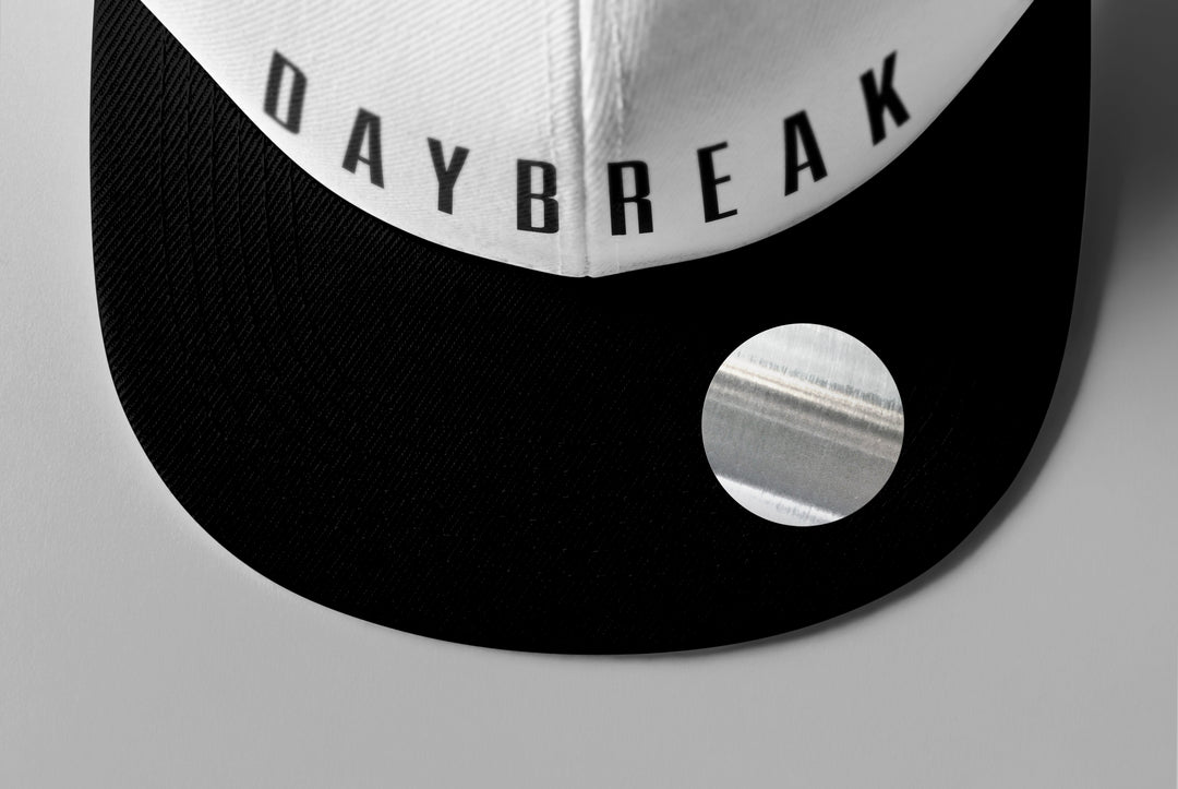 A close-up of a black and white Woe Is Me cap with DAYBREAK printed on the front panel, inspired by the Daybreak album, featuring a shiny circular sticker on the visor.