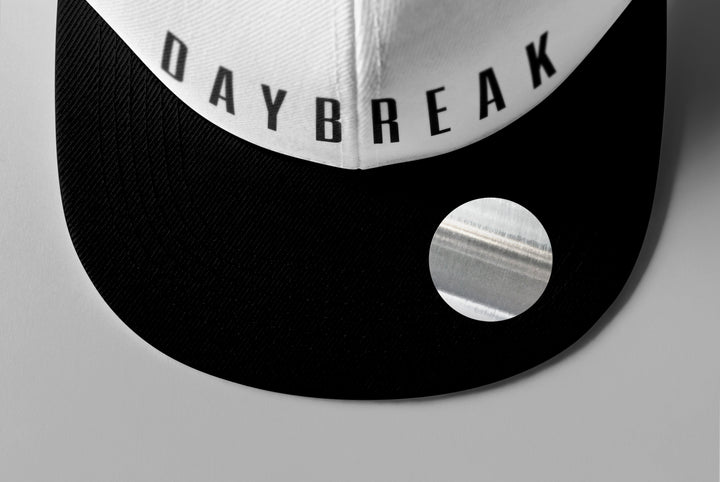 A close-up of a black and white Woe Is Me cap with DAYBREAK printed on the front panel, inspired by the Daybreak album, featuring a shiny circular sticker on the visor.